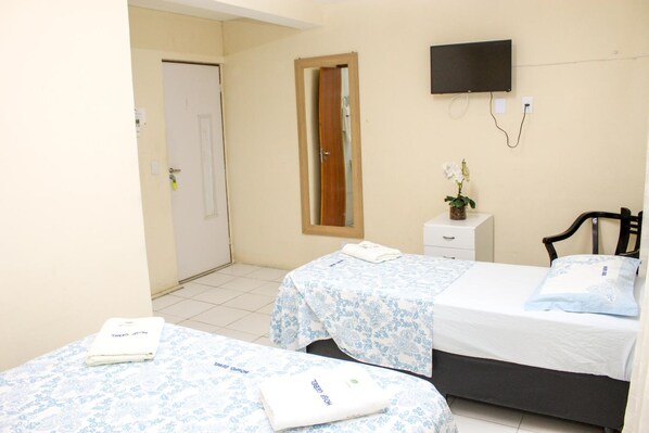 A hotel room with two beds, a TV mounted on the wall, a small bedside table with a vase of flowers, and a door leading to another room.