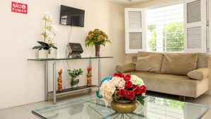 A living room with a glass coffee table, a beige sofa, a TV mounted on the wall, and a window with white shutters. - Hospedaria Gebel LocDay (Recife)