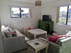 Sala de estar com sofá, dois pufes, mesa de centro, televisão em um armário e janelas amplas com vista para o exterior.