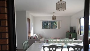 A dining area with a white table and chairs, a hanging light fixture, and a view of greenery outside. - Sunny Holiday Home Near the Beach (Tauranga)