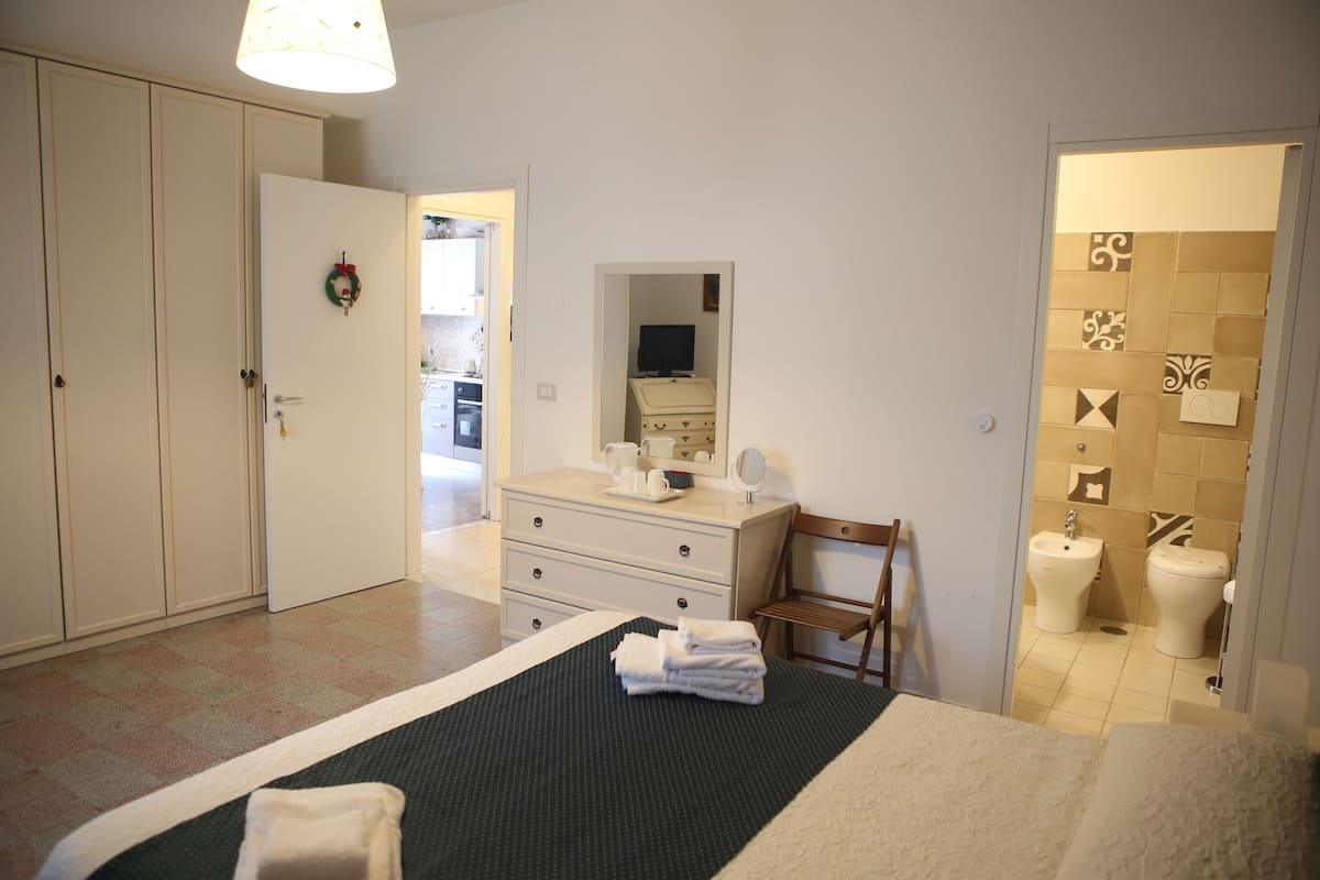 A hotel room with a bed, a dresser, a vanity with a mirror, a chair, and a bathroom with a toilet and bidet.