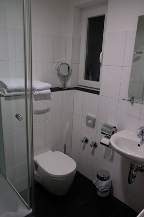 Un baño con inodoro, lavamanos, cabina de ducha con mampara de vidrio y una ventana pequeña.