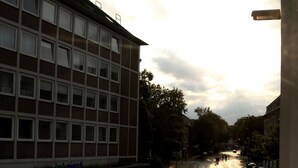 A multi-story building with a row of windows on the ground floor, a visible storefront with signage, and a curved road with lane markings. - Minx - Cityhotels (Aachen)