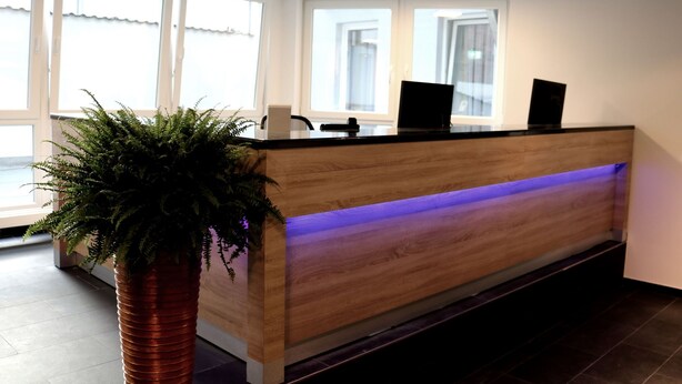 A reception desk with a wooden finish, a potted plant, and a blue-lit reception area. - Minx - Cityhotels (Aachen)
