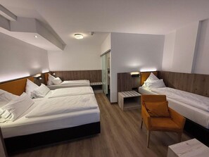 Hotellihuone, jossa on kolme sänkyä, puulattia ja pieni oleskelualue, jossa on yksi tuoli.