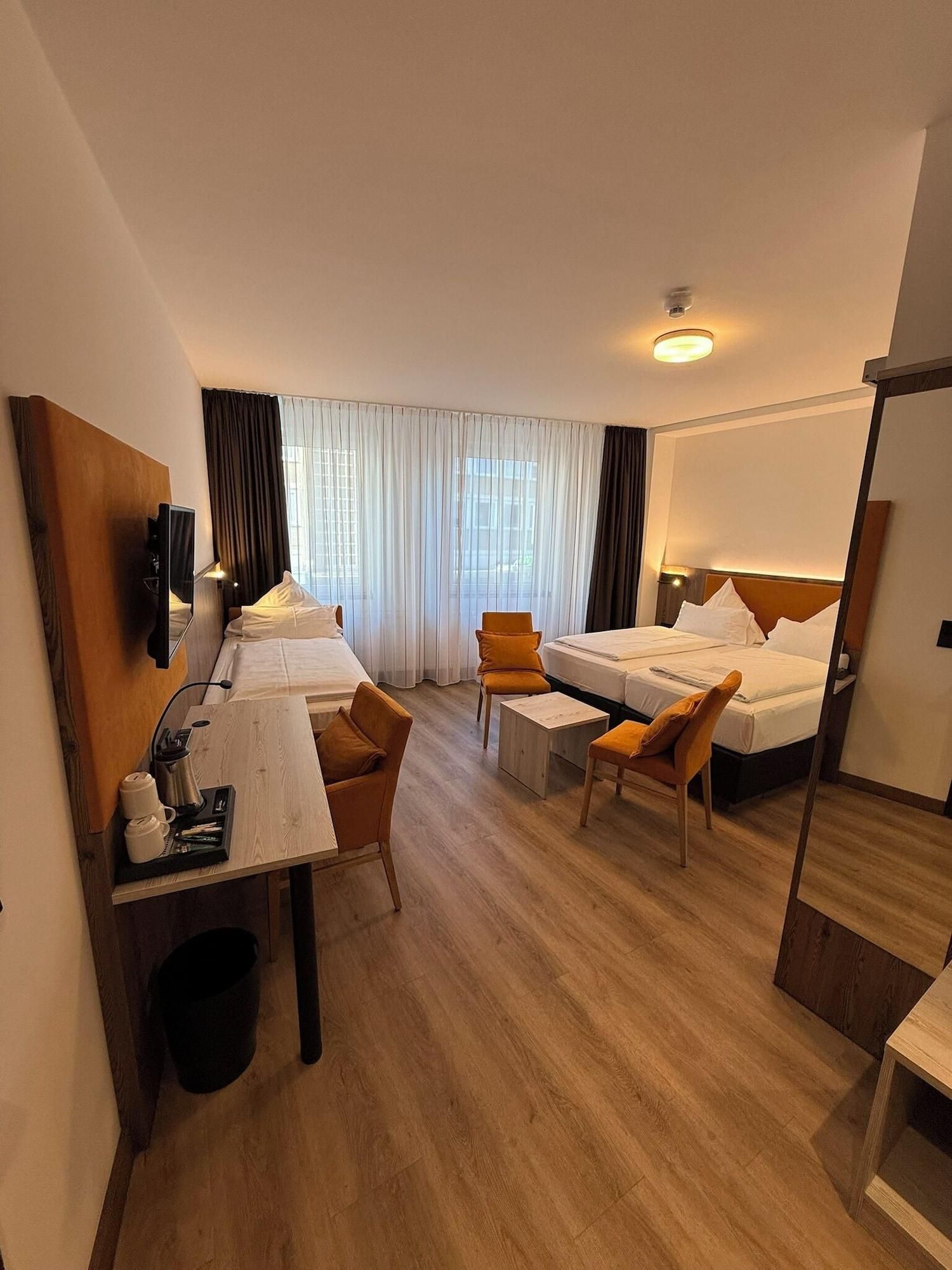 A hotel room with two beds, a desk, chairs, a TV, and a large window with curtains.