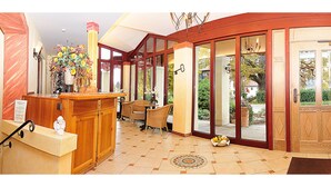 Una hall d'albergo con una reception, sedie in vimini, un vaso di fiori e vista sull'esterno.