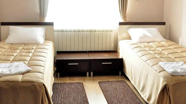 Une chambre d’hôtel avec deux lits, une fenêtre avec des rideaux, un radiateur et une petite table placée entre les lits.