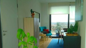 A living room with a potted plant on a table, a blue sofa, a dining table with chairs, and a view through a large window. - Fully Equipped Sunny Apartment (Antwerp)