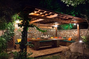 An outdoor patio with a stone wall, wooden pergola, and a table with cushions. - Sapphire Mansion (Karystos)