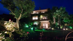 Un bâtiment à deux étages, doté d’un balcon à l’étage supérieur, entouré d’une végétation luxuriante et illuminé par des lampes de jardin.
