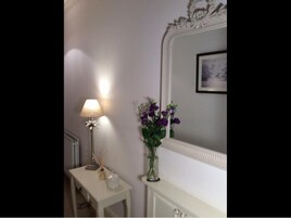 Un tocador blanco con espejo, una mesita con lámpara y un jarrón con flores moradas.