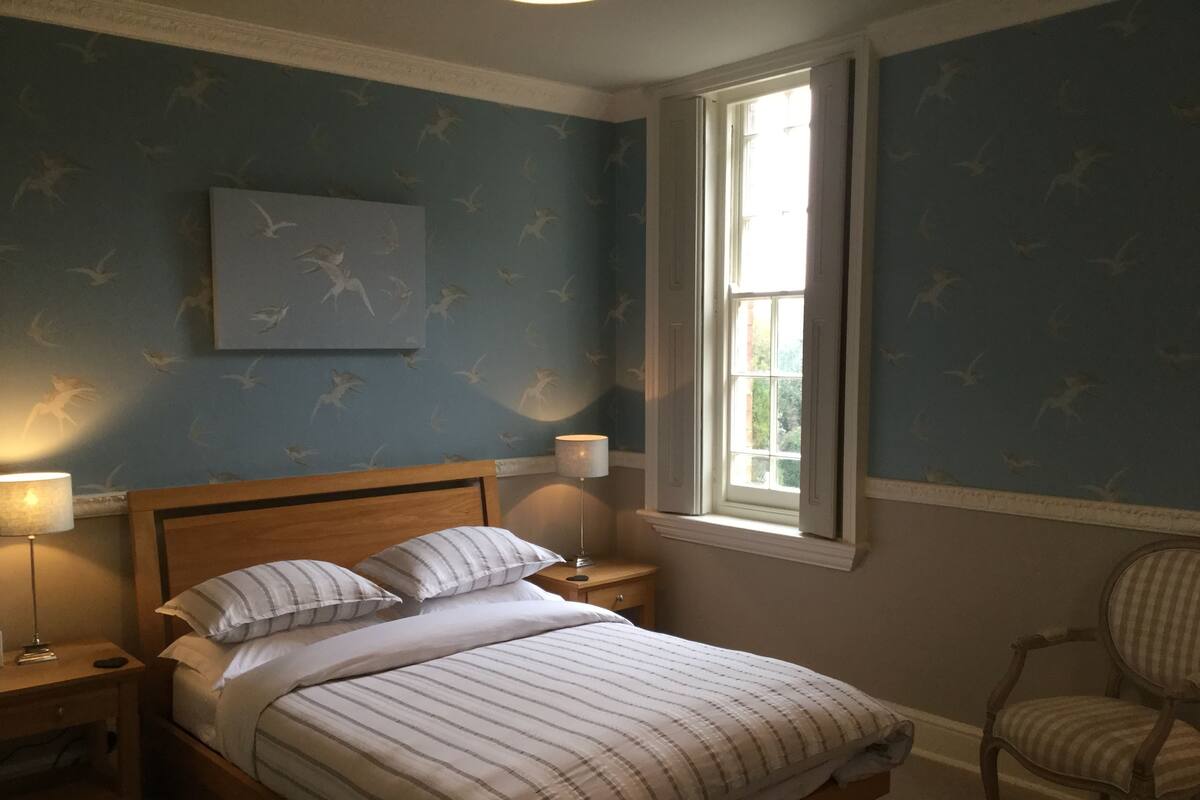 A bedroom with a bed, nightstands, a chair, a window, and a wall with a bird pattern.