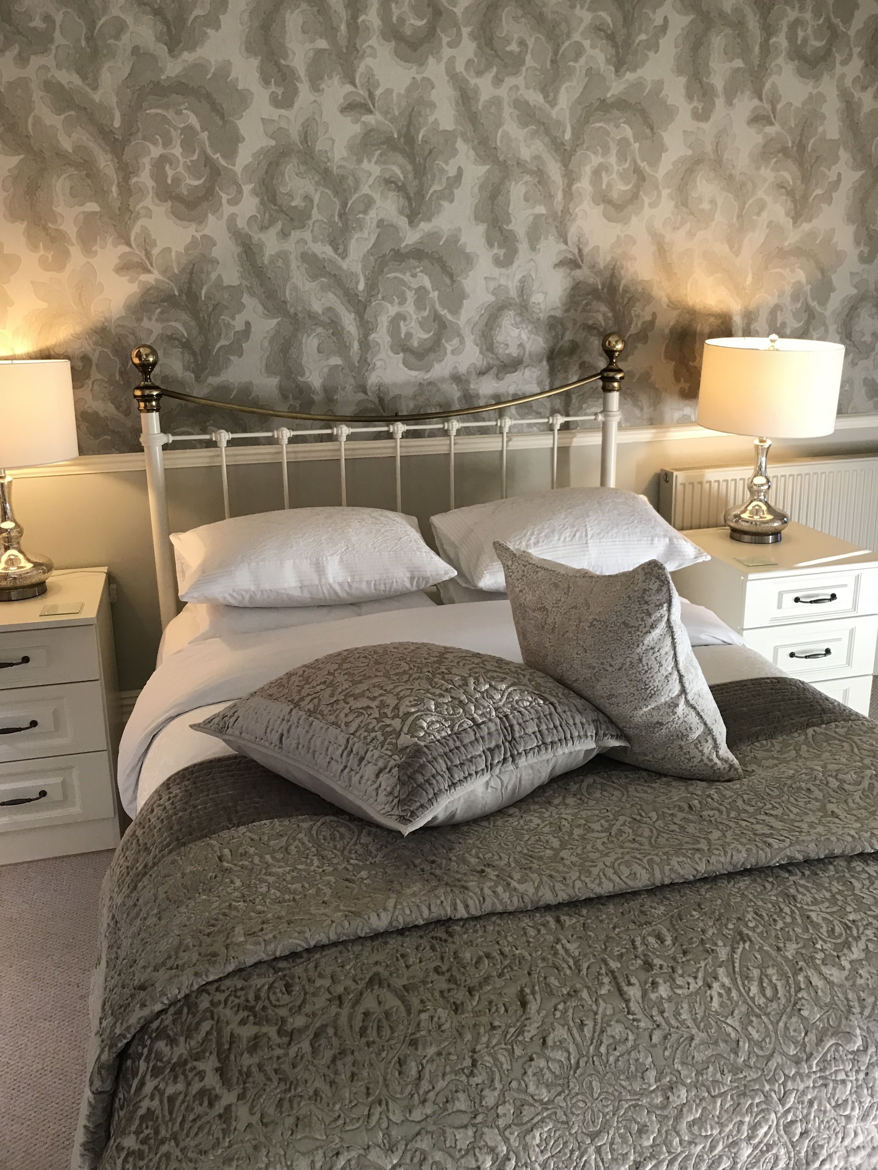 A neatly made bed with a patterned bedspread and two decorative pillows, a white metal headboard, and a wall with a floral wallpaper pattern.