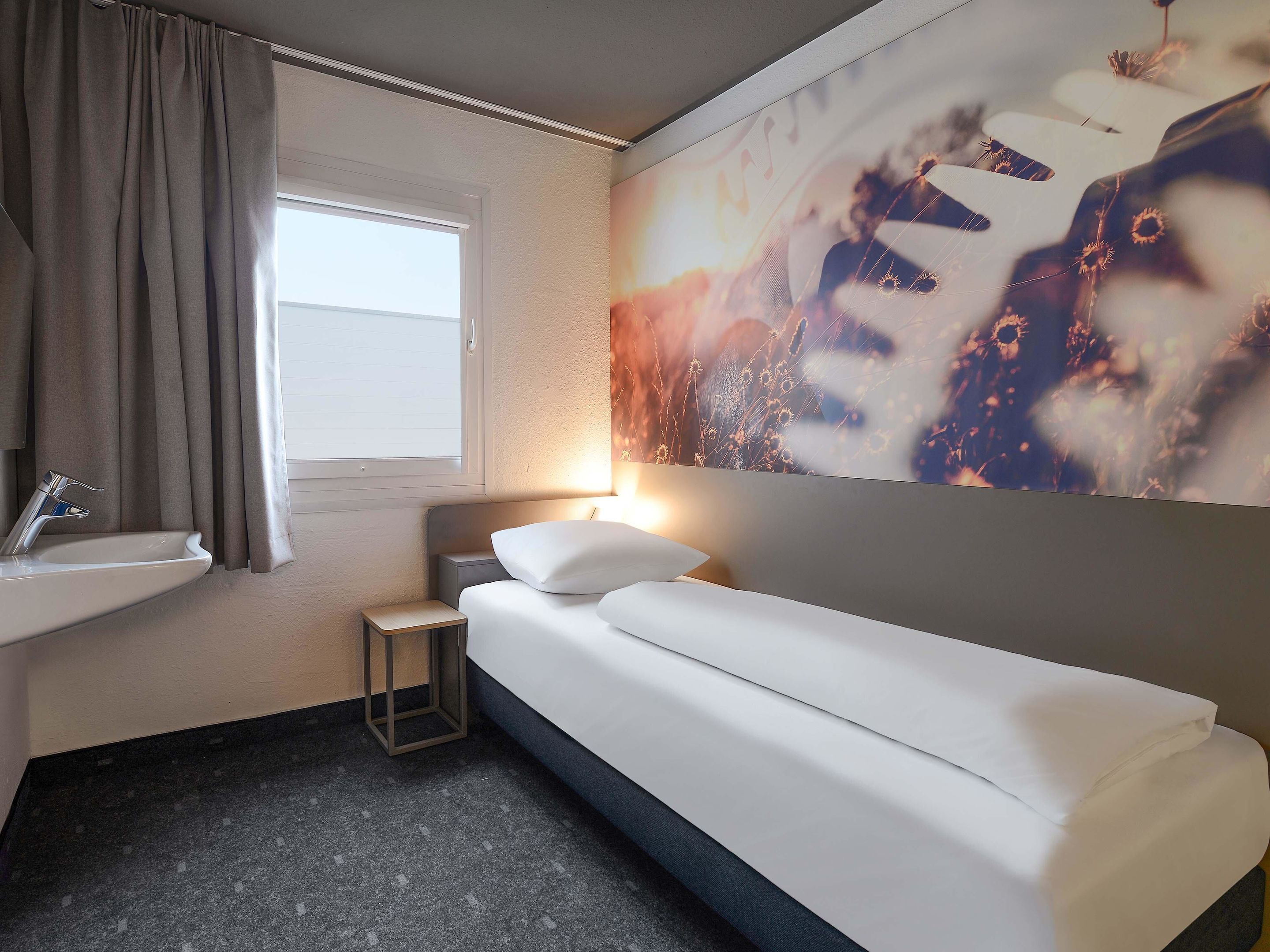 Una habitación de hotel con cama, mesita de noche, lámpara de pared, ventana con cortinas y lavamanos en el baño.