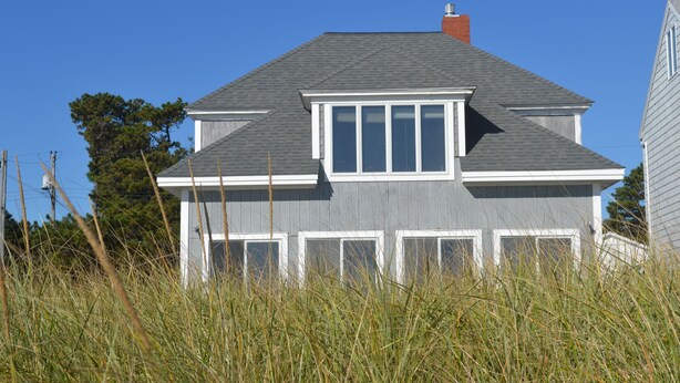 A two-story house with a grey exterior and white trim, surrounded by tall grass. - Sunny Beachfront Cottage with Spectacular Ocean Views  (Saco)