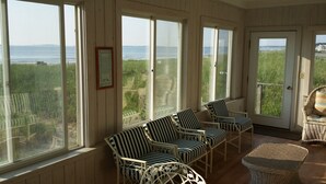 A sunlit room with large windows overlooking a grassy area and the sea. There are wicker chairs and a wicker table. - Sunny Beachfront Cottage with Spectacular Ocean Views  (Saco)