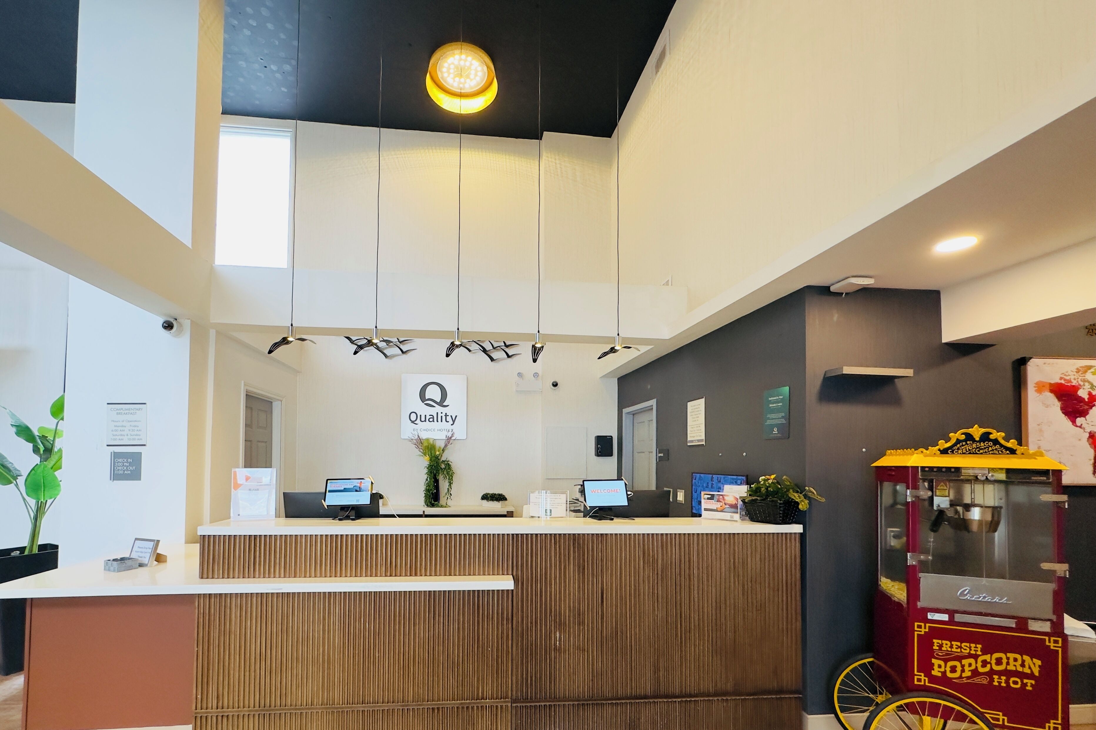 A modern hotel reception area with a wooden reception desk, two computer monitors, a sign reading "Quality," and a popcorn vending machine.