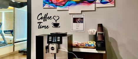 Una estación de café con una cafetera, una pared con un cartel que dice 'Coffee Time' y una cinta de correr al fondo.