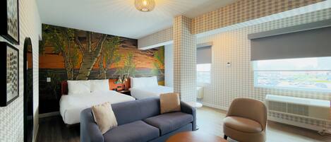 A modern hotel room with a bed, a sofa, a chair, a round ottoman, a mural, and a large window.