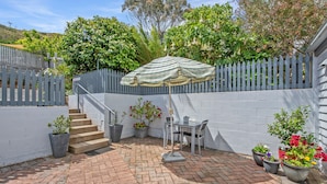 Une terrasse avec une table et des chaises, entourée de plantes en pot et agrémentée d’un parasol à rayures.
