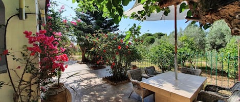 An outdoor patio with a table and chairs, surrounded by potted plants and flowering shrubs.