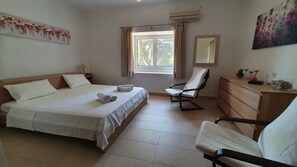 A hotel room with a bed, two chairs, a dresser, a window with curtains, an air conditioner, and artwork on the walls.