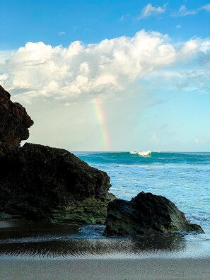 Une plage bordée de formations rocheuses, avec un arc-en-ciel bien visible au-dessus de l’océan.