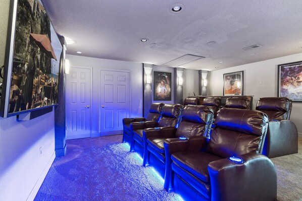 A home theater room with leather reclining chairs, a large flat-screen TV, and wall-mounted posters. - Large outdoor summer kitchen, Private Pool, Theatre Room, 2 miles to Disney ! (Kissimmee)