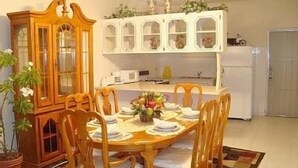 Un coin repas avec une table en bois dressée pour six personnes, entourée de chaises en bois. Un meuble avec des portes en verre est adossé au mur, et un pot de fleurs se trouve sur la gauche.