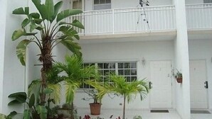 Un bâtiment à deux étages, à la façade blanche, doté d’un balcon à l’étage supérieur et de plusieurs plantes en pot devant l’entrée.