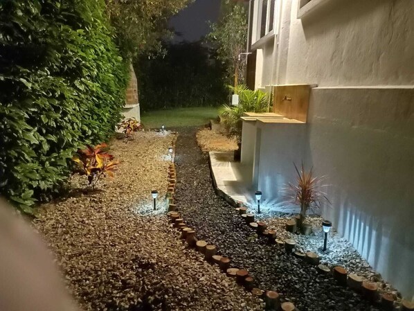 A pathway lined with pebbles and wooden logs, leading to a building with a white exterior and a garden area with plants and a small tree.