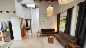 Een ruime woonkamer met een bank, een klein houten tafeltje, een koelkast en een hanglamp.