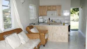 A modern kitchen with wooden cabinets, a white countertop, and a built-in island. There is a wicker sofa with white cushions in the living area.