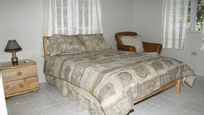 A bedroom with a bed, a nightstand, a lamp, a chair, and a window with sheer curtains.