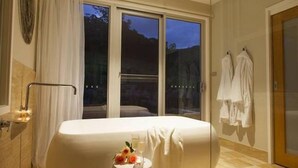 Een moderne hotelkamer met een groot raam, een badkuip en een klein tafeltje met een glas water en bloemen.