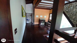 Ein Flur mit Steinwand, Holzbalken und Kamin. An der Wand befinden sich Bilder in Rahmen, und zur Rechten führt eine Treppe.