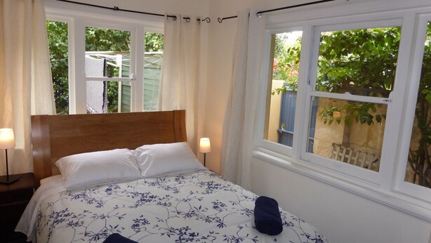 Een slaapkamer met een houten hoofdeinde, een bloemenbedekker, twee bedlampjes en grote ramen met gordijnen.