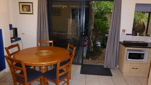 Een knus, klein vertrek met een ronde houten eettafel en vier stoelen, een kitchenette met een magnetron, en een schuifdeur die leidt naar een tuin.