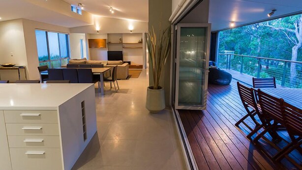 A modern kitchen with a central island, a dining area with a wooden table and chairs, and a balcony with a view of trees. - Flutes Escape - Located on the exclusive Brookland Valley Vineyard property (Wilyabrup)