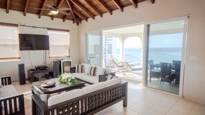 Ampla sala de estar com sofá, mesa de centro e TV, conectada a uma varanda com vista para o mar.