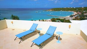 Terraço com duas espreguiçadeiras azuis e uma pequena mesa, com vista para uma praia com barcos e um mar azul límpido.