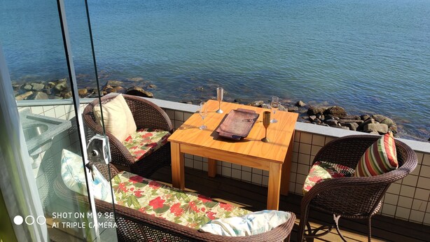 Un balcon agrémenté de meubles en osier, d’une table en bois et offrant une vue sur des bateaux voguant sur une mer calme, avec des montagnes en toile de fond.
