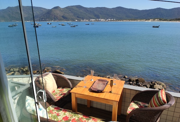 A balcony with wicker furniture, a wooden table, and a view of boats on a calm sea with mountains in the background.