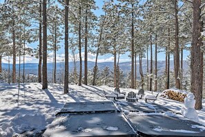 A snow-covered outdoor area with a table and chairs, surrounded by tall trees and a view of distant mountains. - Custom Angel Fire Home: Views, Hot Tub & Fire Pit! (Angel Fire)