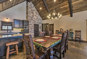 A rustic dining area with a long wooden table set for six, surrounded by wooden chairs. A stone fireplace and a chandelier are visible in the background. - Custom Angel Fire Home: Views, Hot Tub & Fire Pit! (Angel Fire)