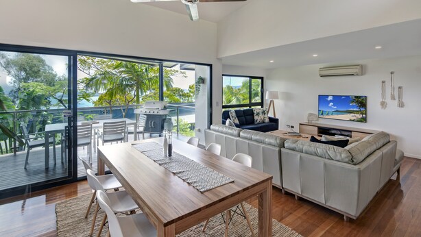 Een moderne woonkamer met een houten eettafel, witte stoelen, een grijze bank, een televisie en een balkon met uitzicht op de oceaan.