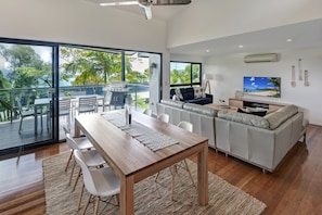 A modern living room with a wooden dining table, white chairs, a grey sofa, a TV, and a balcony with a view of the ocean.