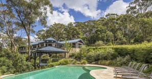A pool area with a curved edge, surrounded by lush greenery and a large house with a covered patio in the background.