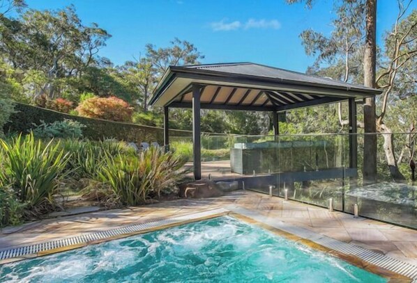 A swimming pool with a glass enclosure, surrounded by lush greenery and a covered seating area.